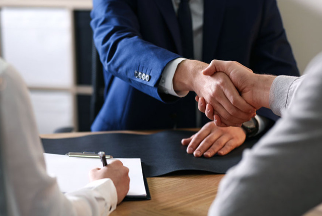 Imagen que muestra unas manos estrechándose como que están llegando a un acuerdo entrea las partes, otra persona está formando un contrato